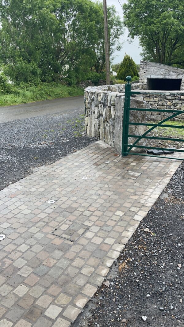 A stone wall and a green metal gate mark the entrance to a gravel road, with a patterned stone pathway leading up to the gate, surrounded by trees and grass. The scene is wet, suggesting recent rain.