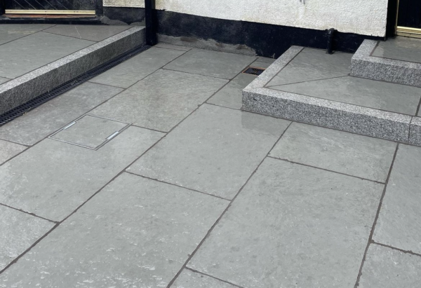 A finished patio area with gray stone tiles, featuring steps leading to the entrance of a house. A drain cover is visible, and a wall displays a decorative wind chime. The setting appears modern and well-maintained.