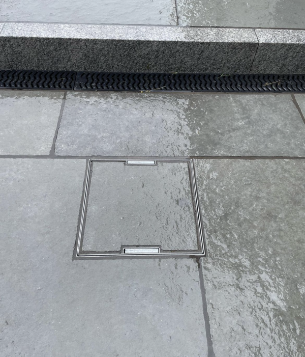 A close-up of a rectangular metal access cover set into a wet stone pavement, surrounded by grey stone tiles, with a textured black drainage strip nearby.