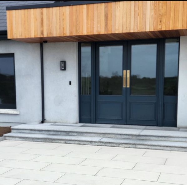 A modern entrance featuring two large blue doors with golden handles, a wooden overhang, and stone steps leading up to the doorway, set against a light gray exterior wall.