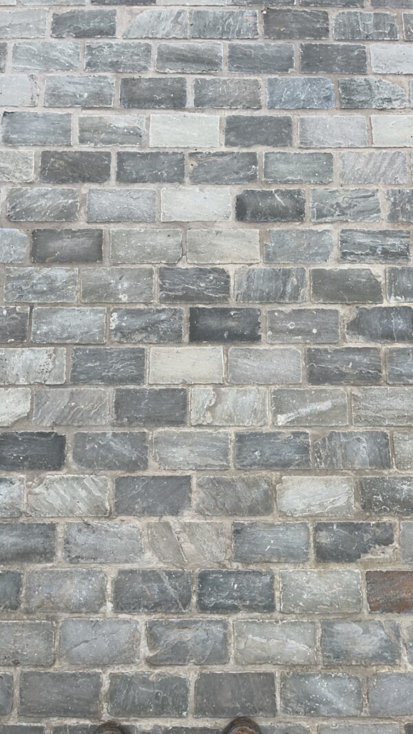 A close-up view of a patterned stone pavement made of various shades of gray and blue stones, with visible joints and some wear, and a pair of brown boots at the bottom of the image.