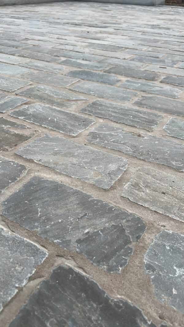 A close-up view of unevenly laid gray stone pavers with visible joints filled with sand.