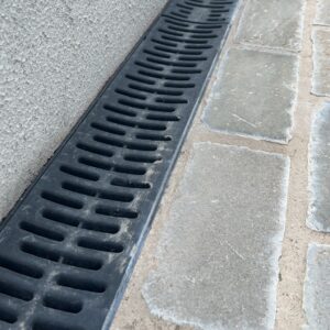 A close-up of a black drainage grate positioned along a textured wall, alongside cobblestone paving.