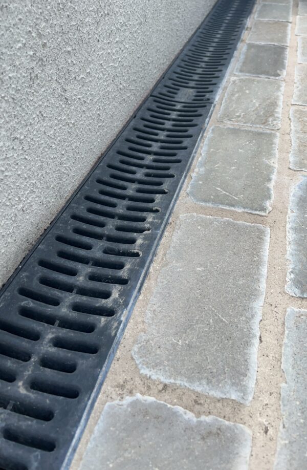 A close-up of a black drainage grate positioned along a textured wall, alongside cobblestone paving.