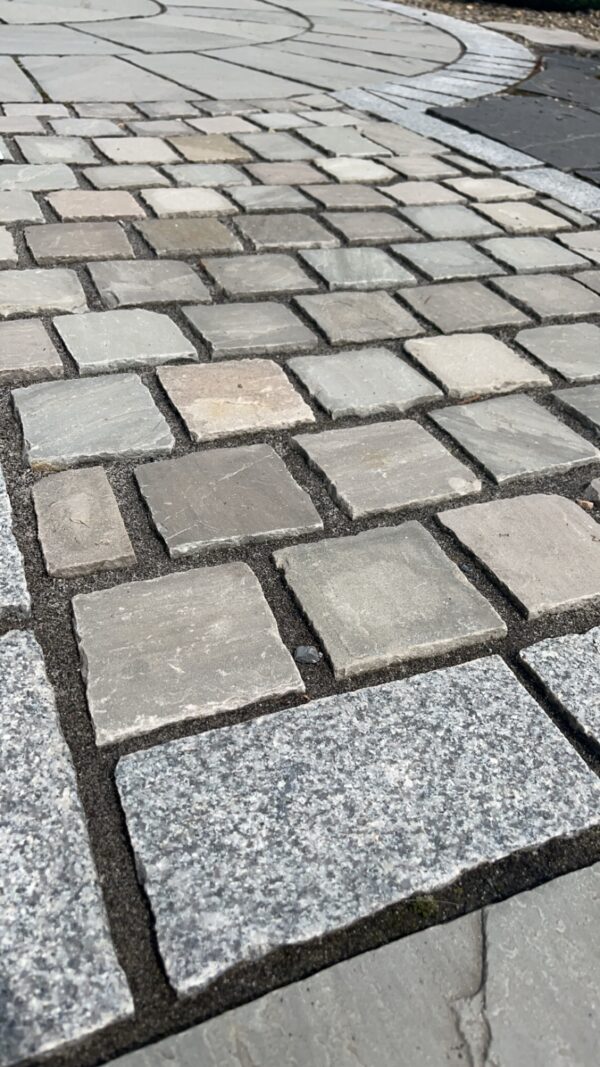 Close-up of a patterned stone pavement featuring a mix of square and rectangular stones in various shades of gray, beige, and light brown, with visible joints filled with dark sand.