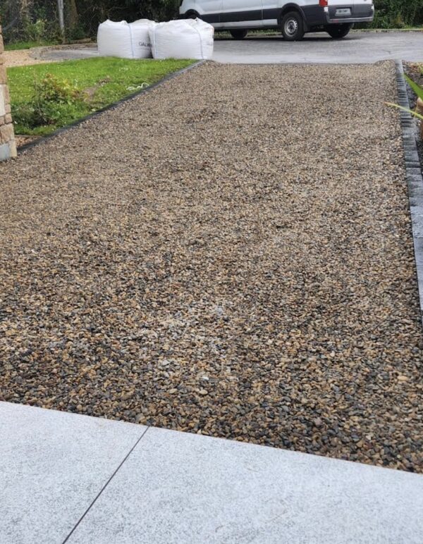 A gravel driveway with a smooth concrete edge and bags of gravel in the background, leading towards a parked vehicle.