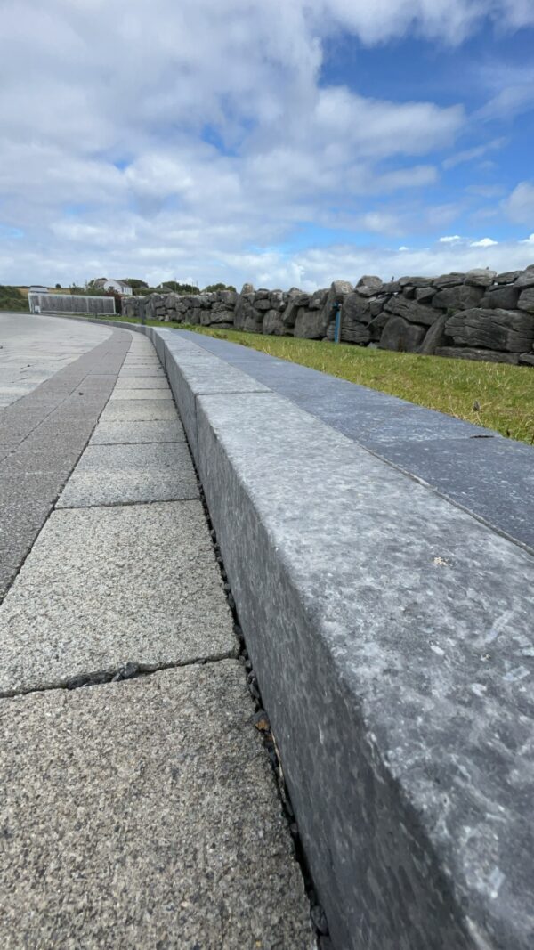 A close-up view of a gray stone edge beside a walkway, with a grassy area and a rocky wall in the background under a cloudy sky.