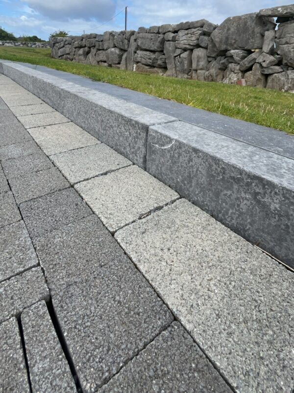 A close-up view of a stone pathway made of rectangular gray and beige tiles, with a grassy area and a low stone wall in the background under a partly cloudy sky.