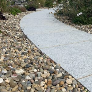 A winding gray stone pathway bordered by colorful pebbles, surrounded by lush green plants and flowers under a blue sky.