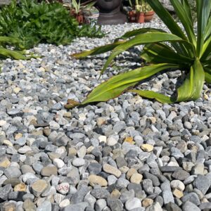 A serene garden scene featuring a large stone Buddha head partially obscured by greenery, surrounded by a bed of small gray and beige pebbles, with a textured wooden border in the foreground and a stone wall in the background.