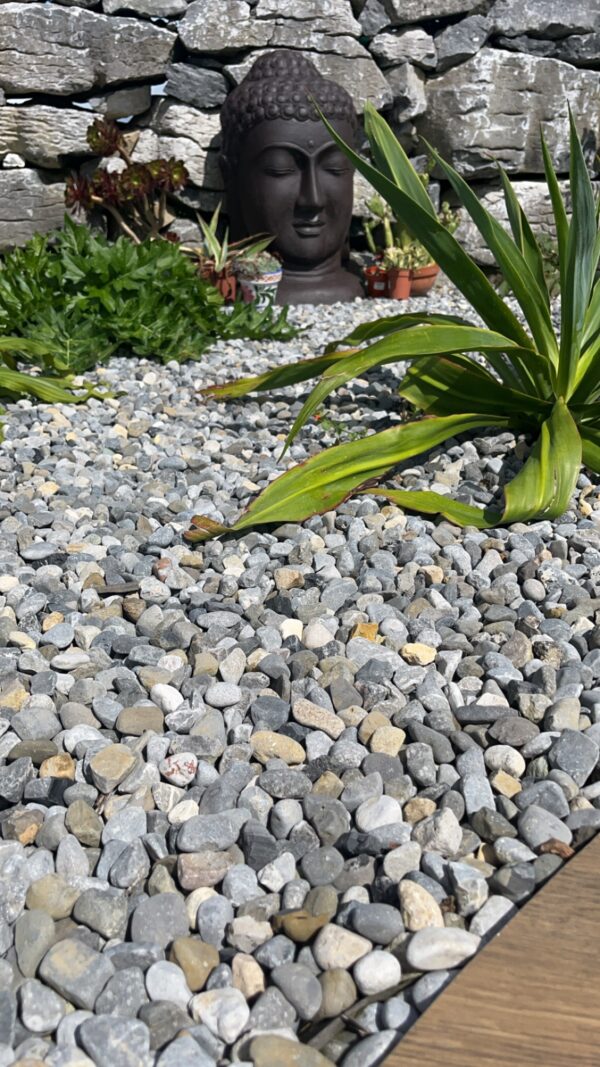 A serene garden scene featuring a large stone Buddha head partially obscured by greenery, surrounded by a bed of small gray and beige pebbles, with a textured wooden border in the foreground and a stone wall in the background.