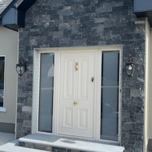 A modern front entrance featuring a white door with decorative panels, flanked by large windows, and surrounded by dark stone cladding. Two lantern-style light fixtures are mounted on either side of the door, with a short set of gray stone steps leading up to the entryway.