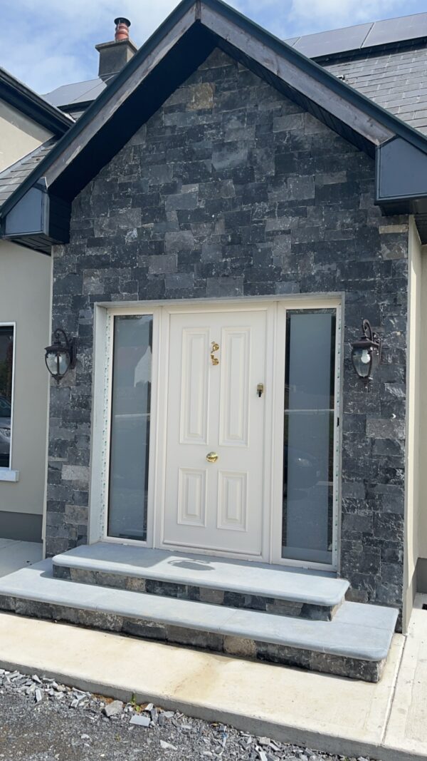 A modern front entrance featuring a white door with decorative panels, flanked by large windows, and surrounded by dark stone cladding. Two lantern-style light fixtures are mounted on either side of the door, with a short set of gray stone steps leading up to the entryway.