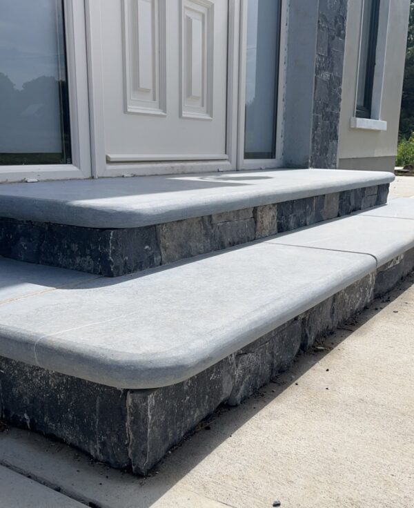 A close-up view of a concrete entrance with three steps, featuring a smooth gray finish and stone accents, leading up to a white door with a brass doorknob.