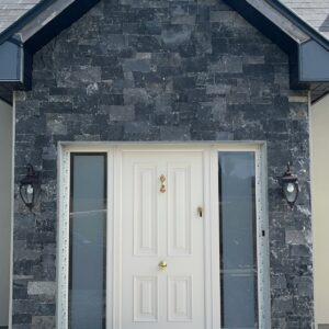 Front entrance of a modern house featuring a symmetrical design with a white double door, flanked by two vertical windows, and a stone facade in shades of gray. The entrance includes a wide stone staircase and decorative lanterns on either side. Solar panels are visible on the roof above.