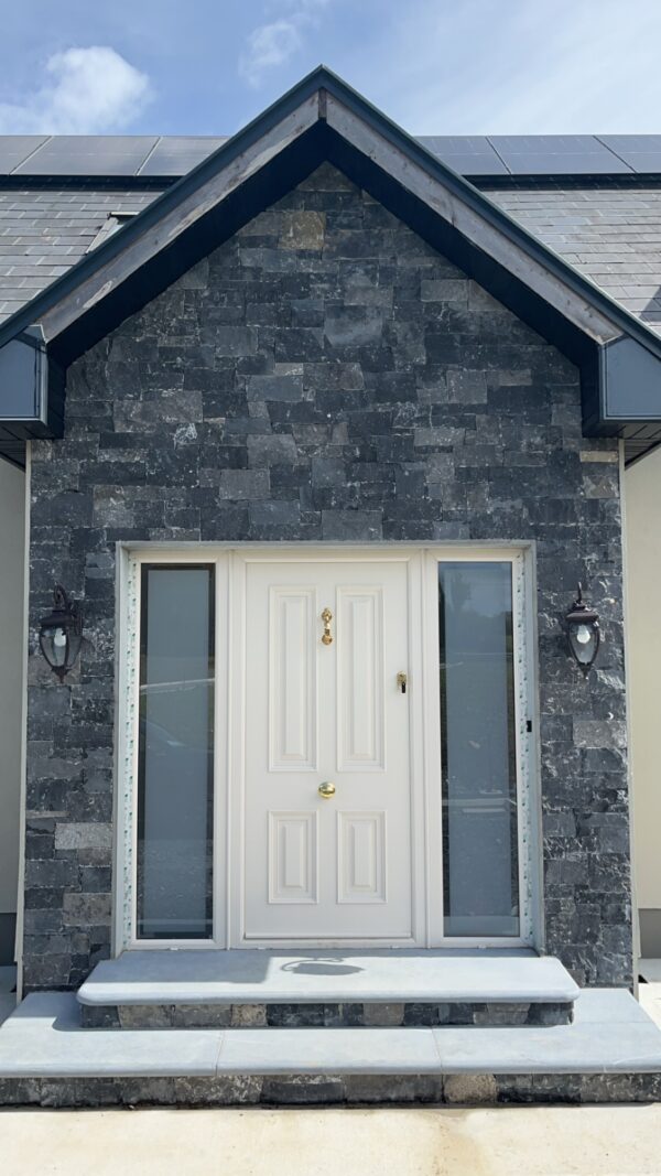 Front entrance of a modern house featuring a symmetrical design with a white double door, flanked by two vertical windows, and a stone facade in shades of gray. The entrance includes a wide stone staircase and decorative lanterns on either side. Solar panels are visible on the roof above.