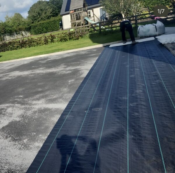 A person is laying down a long black tarpaulin with green lines on a gravel surface in a yard. In the background, a house with a solar panel and a children's playset is visible, surrounded by greenery.