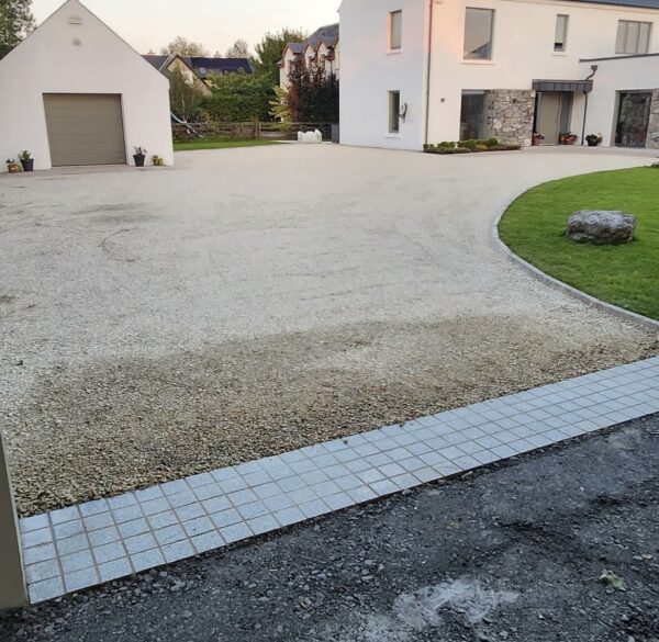 A view of a residential driveway featuring a gravel surface with a paved edge, leading to a modern white house with large windows and a stone façade. A small garage is visible on the left, and green grass and trees can be seen in the background.