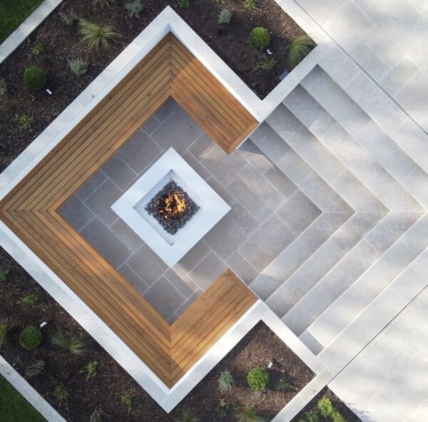 Aerial view of a modern outdoor fire pit surrounded by angular wooden seating and patio stones, with landscaped plants in the background.