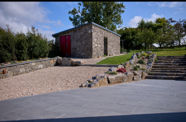 A modern stone building with red double doors, surrounded by a landscaped area featuring a gravel pathway, stone walls, and steps leading up to a lush green garden under a partly cloudy sky.