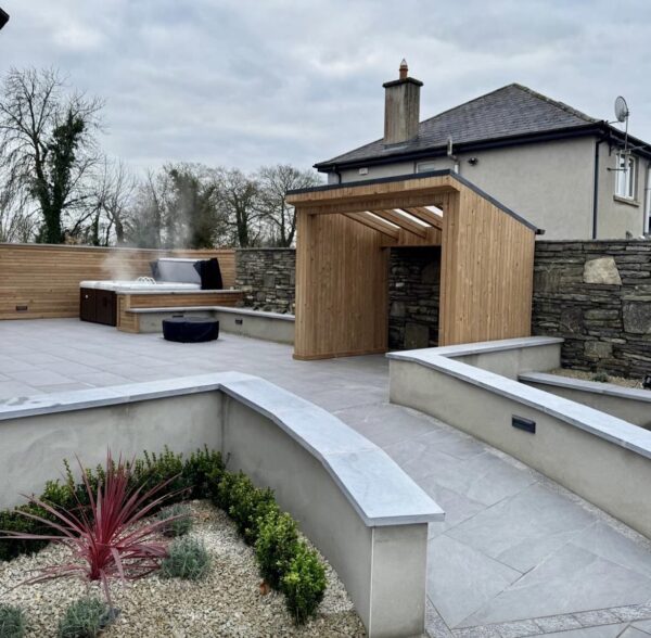 A modern outdoor patio area featuring a wooden gazebo, a hot tub emitting steam, stone walls, and landscaped gardens with plants and gravel paths under a cloudy sky.