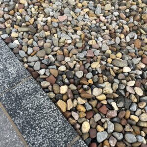 A close-up view of a textured surface made of various small, rounded pebbles in shades of gray, brown, and red, bordered by a dark granite edge.