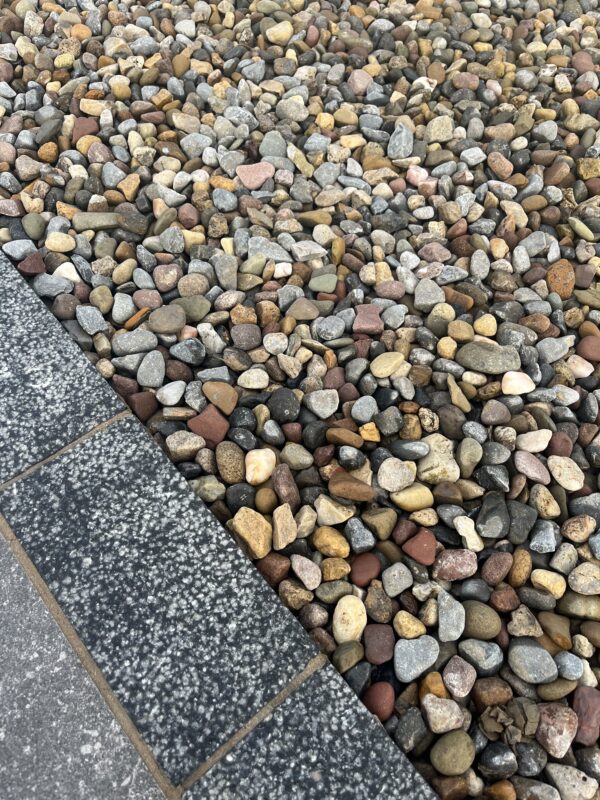 A close-up view of a textured surface made of various small, rounded pebbles in shades of gray, brown, and red, bordered by a dark granite edge.