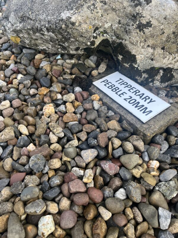 A close-up view of various small pebbles in different colors, with a large rock in the background and a sign labeled "TIPPERARY PEBBLE 20MM" partially visible on the ground.