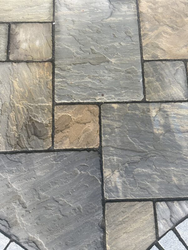 Close-up view of a textured stone paving surface featuring various shades of gray, beige, and brown, arranged in an irregular pattern with dark grout lines.