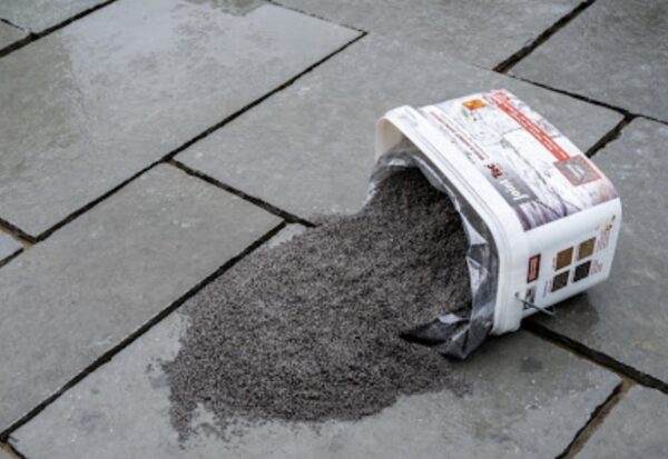 A spilled bucket of gray paving jointing material on wet stone paving slabs.