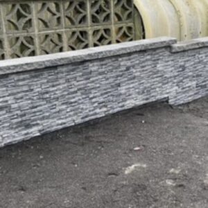 A long, low stone wall made of stacked gray stones, with a decorative patterned wall visible behind it. The ground is sandy and dark.