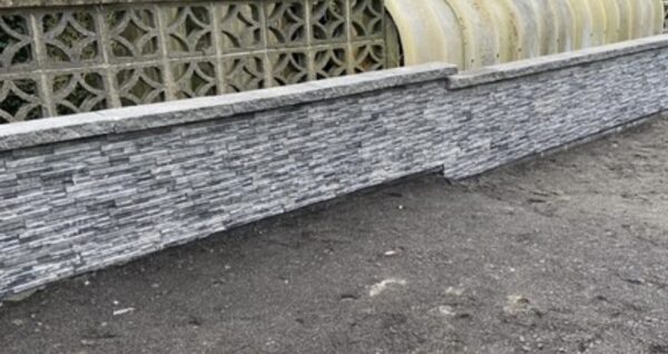 A long, low stone wall made of stacked gray stones, with a decorative patterned wall visible behind it. The ground is sandy and dark.