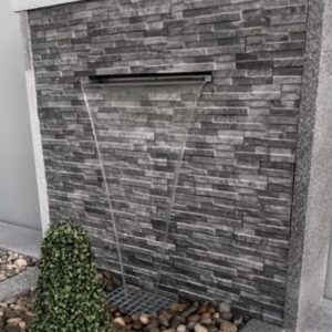 A modern outdoor space featuring a stone wall fountain, two conical topiary shrubs, and a bed of smooth pebbles, all set against a neutral background.