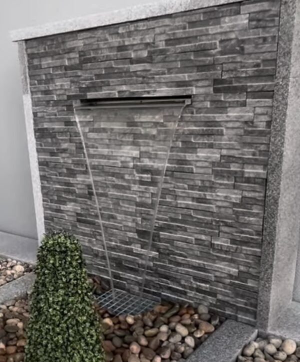 A modern outdoor space featuring a stone wall fountain, two conical topiary shrubs, and a bed of smooth pebbles, all set against a neutral background.