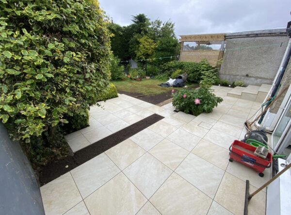 A landscaped garden area featuring light-colored tile flooring, a border of dark soil, lush green hedges, and scattered flower pots, with a small lawn and garden tools visible in the background.