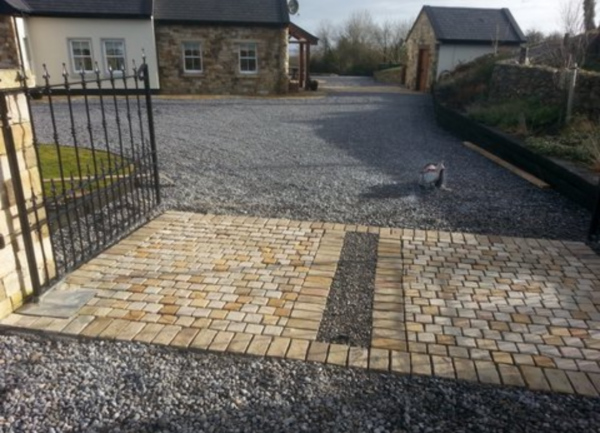 A gravel driveway with a gated entrance, featuring a patterned stone surface leading into a spacious yard and two nearby buildings, one of which has a stone facade.