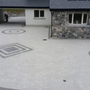 A modern house with a stone and light-colored exterior, featuring a patterned driveway with circular and square designs. A person stands near an entrance, and there are several windows visible.