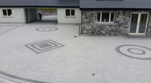 A modern house with a stone and light-colored exterior, featuring a patterned driveway with circular and square designs. A person stands near an entrance, and there are several windows visible.