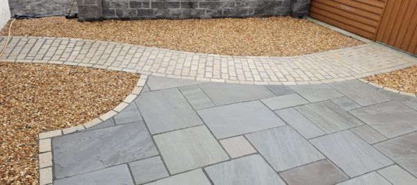 A landscaped outdoor area featuring a stone wall, wooden fence, and a mix of gravel and paved paths in shades of grey and beige. A ladder leans against a textured wall in the background.