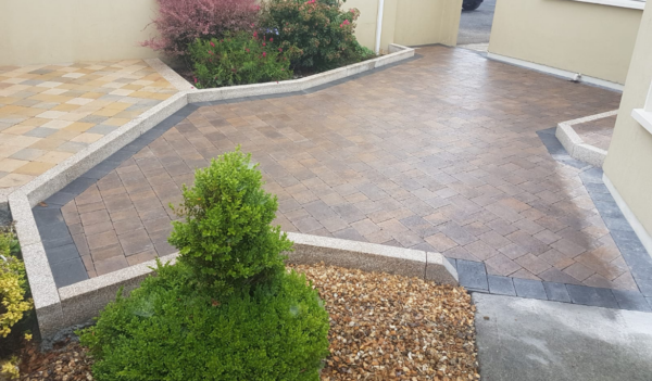 A landscaped outdoor area featuring patterned paving stones, bordered by a low stone wall, with colorful shrubs and a rounded green bush, and a house exterior in the background.