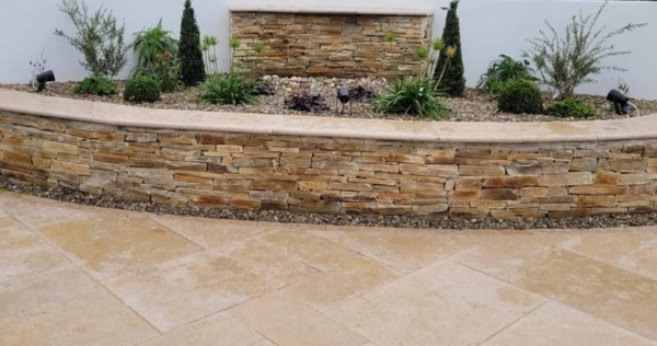 A landscaped garden featuring a curved wall made of Egyptian limestone, adorned with various plants and shrubs, set against a white background.